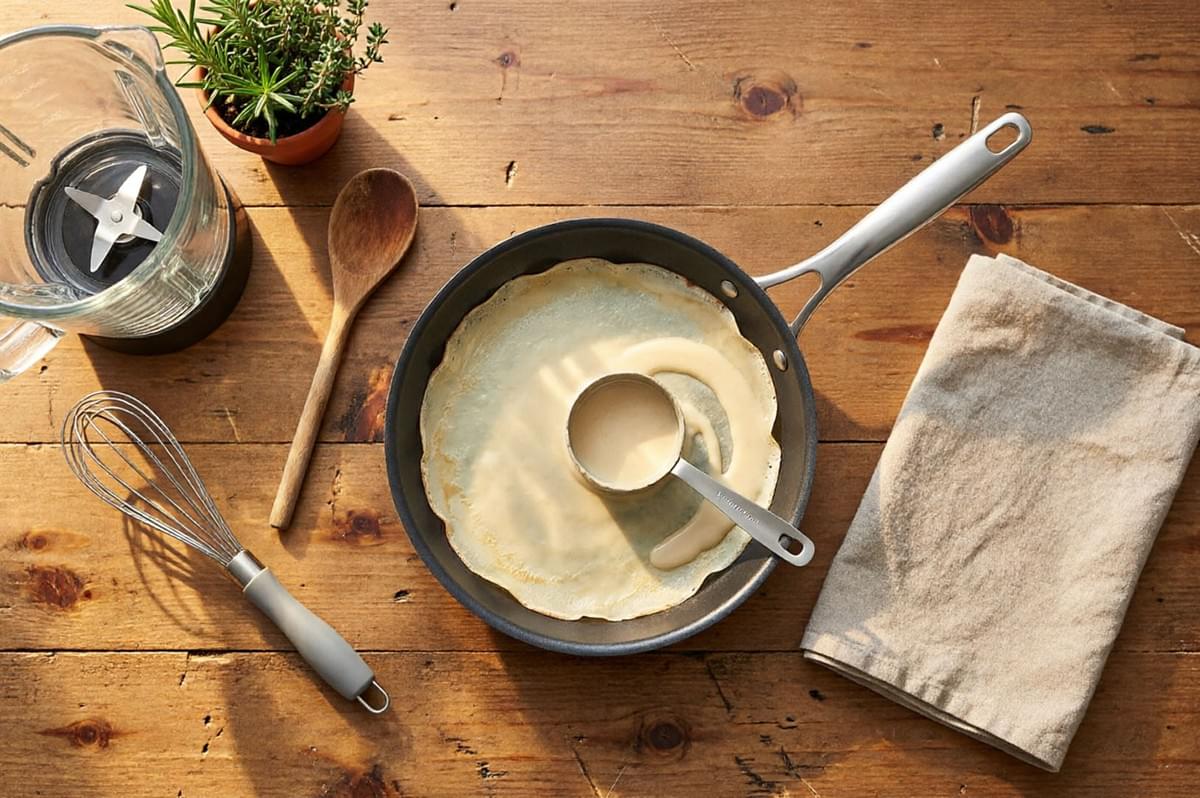 Overhead view of crepe batter being poured into pan with whisk, blender, and fresh herbs on wooden table