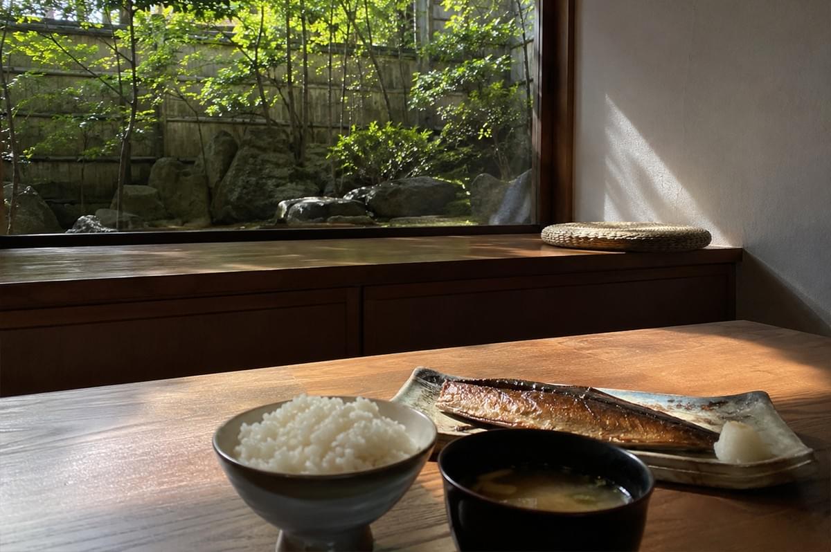 A traditional Japanese meal is neatly arranged on a wooden dining table, illuminated by a warm beam of natural sunlight. In the foreground, the set features a ceramic bowl of fluffy white rice, a dark lacquer bowl filled with miso soup, and a rustic rectangular plate holding a piece of grilled fish with crispy, glistening skin alongside a small dollop of grated radish. Behind the table, a wide wooden ledge runs along a large glass window where a circular woven straw cushion rests. The window looks out into a serene, sun-drenched Japanese courtyard garden complete with lush green trees, ornamental shrubs, large mossy rocks, and a natural bamboo fence, establishing a peaceful and authentic dining ambiance.