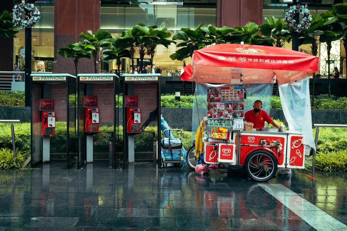 An ice cream vendor in a red shirt stands next to his covered Wall's cart on a wet city sidewalk, with a row of three public payphone booths to his left. In the background, green palm plants and decorated pillars frame a modern storefront reflection on the ground.