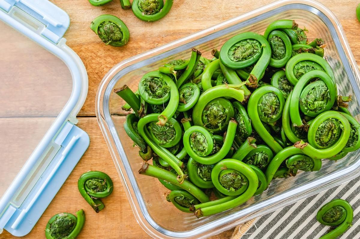 A stack of six lemon bars on a white square plate, showcasing their vibrant yellow filling. A large, juicy lemon wedge sits in the foreground against a light green textured background, emphasizing the fresh, citrusy ingredients.