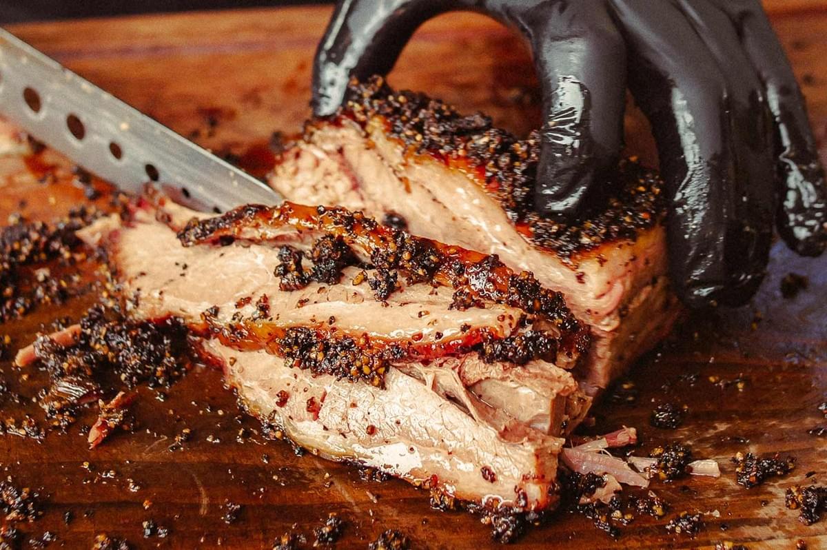 Close-up of a smoked brisket with a dark pepper crust being sliced on a wooden board by a gloved hand, showing juicy, marbled interior and a sharp knife.