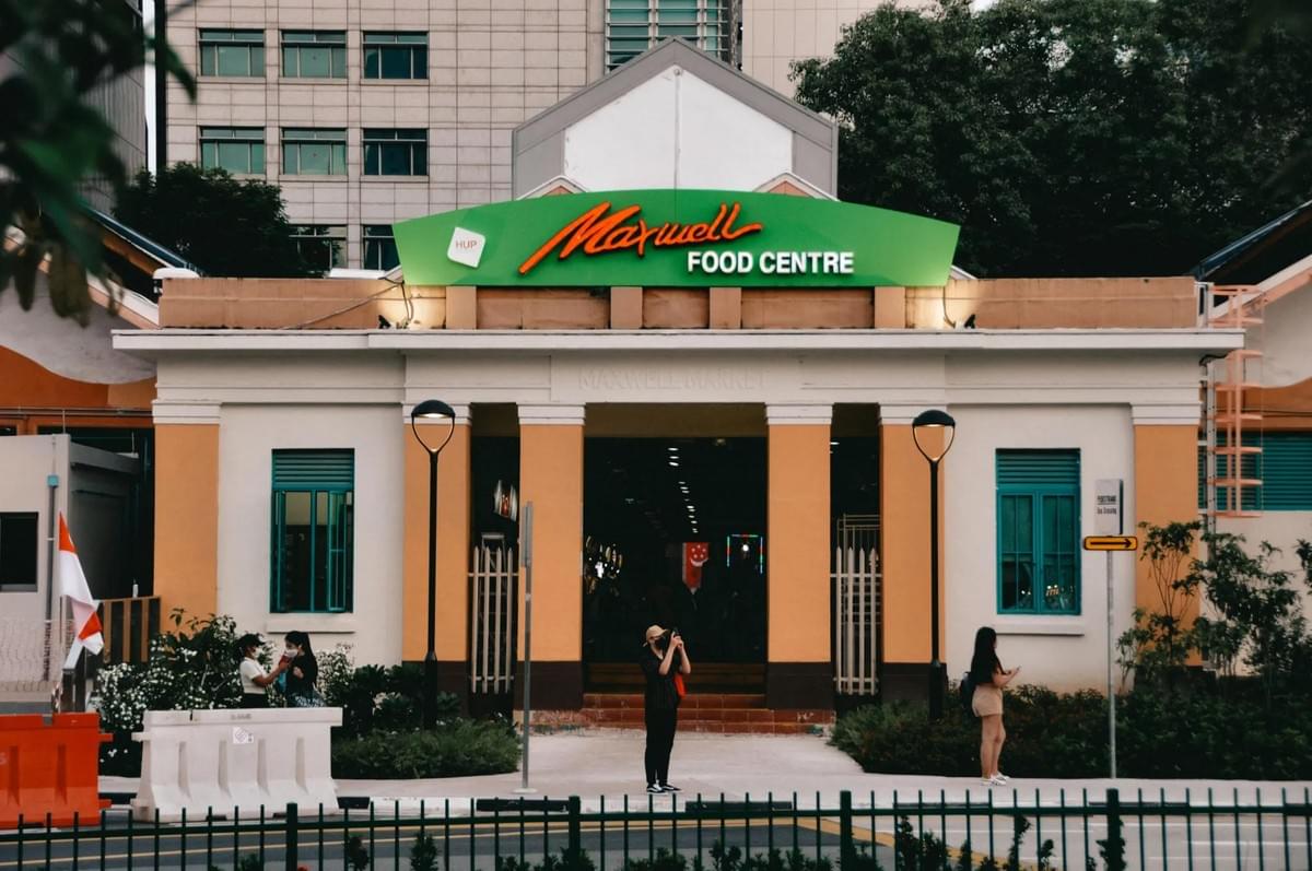 The exterior entrance of the Maxwell Food Centre features a large green sign with orange script lettering above an open, arched doorway. Pedestrians stand on the sidewalk in front of the beige and orange building, which is framed by green windows and a black fence.