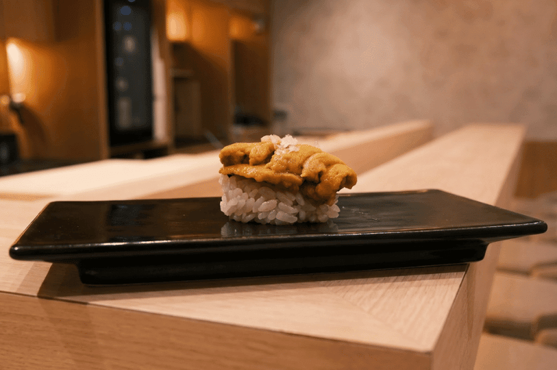 A macro close-up of a fresh uni nigiri sushi piece topped with flaky sea salt, resting on a glossy black plate at Sushi Masa by Ki-setsu.