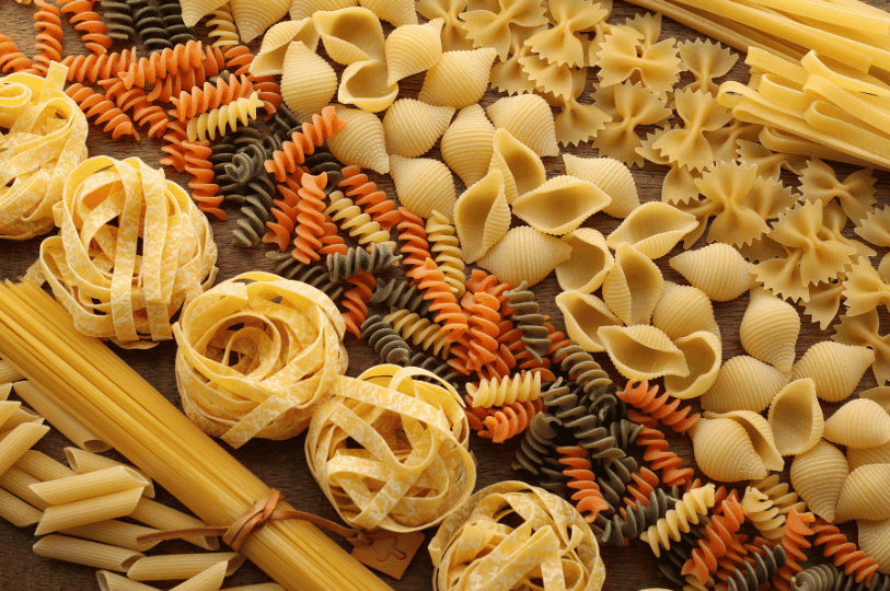 A vibrant, textured display of mixed Italian pasta shapes arranged in diagonal rows, including bow-tie farfalle, ridged shells, tricolor fusilli, and ribbon pasta.