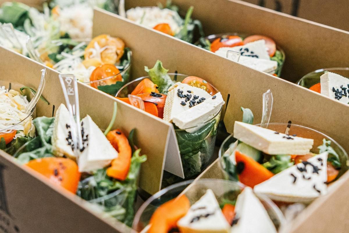 This image shows several small salad cups neatly arranged inside a cardboard catering box with dividers. Each portion features fresh greens, sliced tomatoes, and a wedge of tofu topped with black sesame seeds.