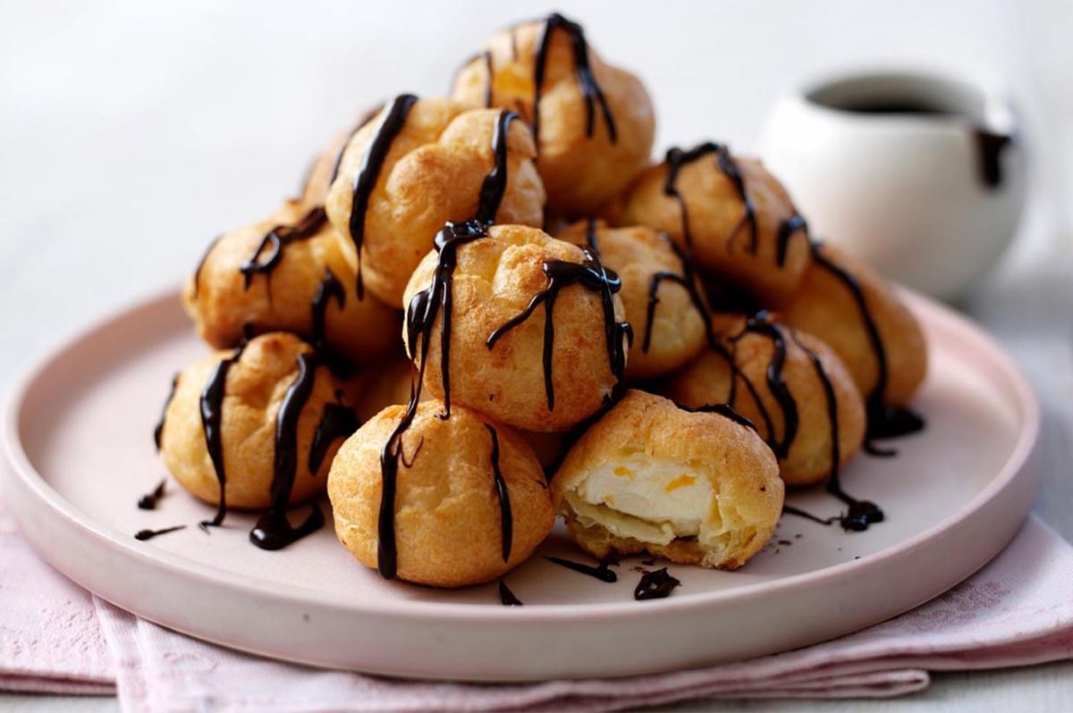 A tall stack of profiteroles (cream puffs filled with white cream) drizzled with dark chocolate sauce, served on a light pink plate. One puff is cut open to show the filling.