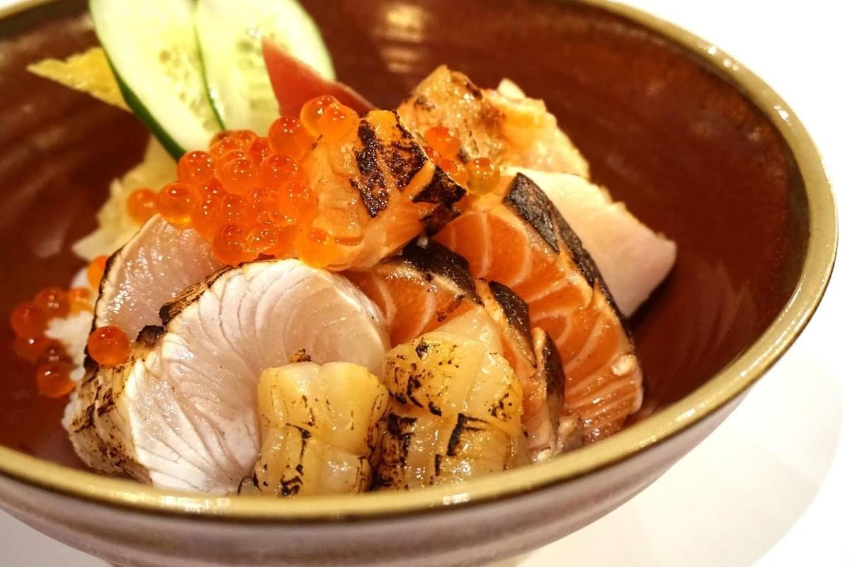 A brown ceramic bowl filled with a colorful Japanese chirashi don dish featuring seared fish slices, scallops, and salmon roe. Cucumber slices and ginger garnish the edge of the bowl above the seafood.