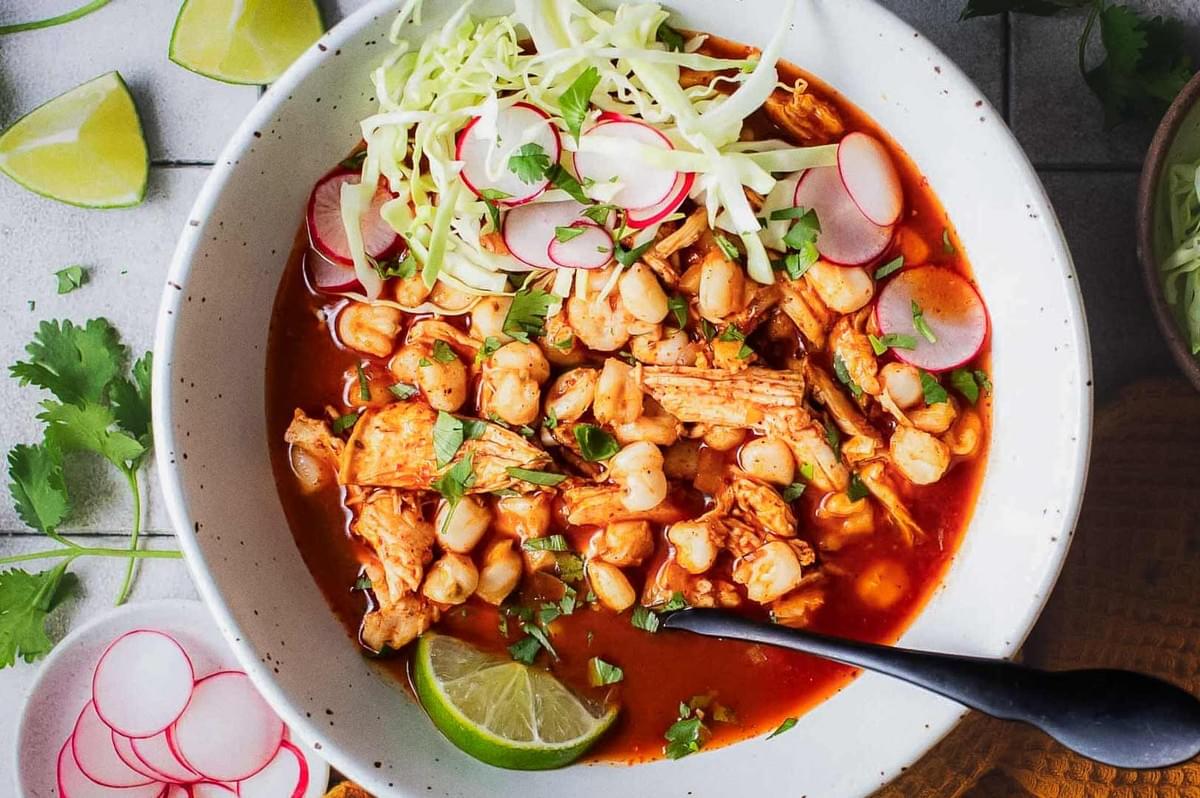 Mexican pozole with chicken in red chili broth, garnished with cabbage, radishes, cilantro, and lime, traditional hominy soup presentation.