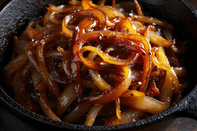 A close-up shot of golden-brown caramelized onion ribbons in a seasoned cast-iron skillet, showing a glossy, jam-like texture and rich color.