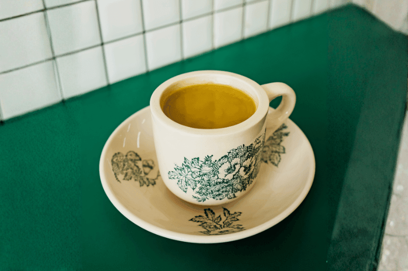 A serving of milky yellow-brown Singapore kopi in a classic green floral kopitiam cup and saucer, placed on a bright green counter in front of a white square-tiled wall.