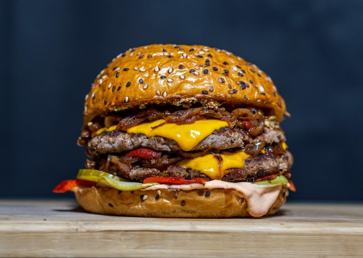 A gourmet double cheeseburger stacked with two beef patties, melted cheese, and caramelized onions sits on a wooden board against a dark background. The toasted sesame seed bun holds layers of sliced pickles, red peppers, and creamy sauce dripping along the bottom edge.