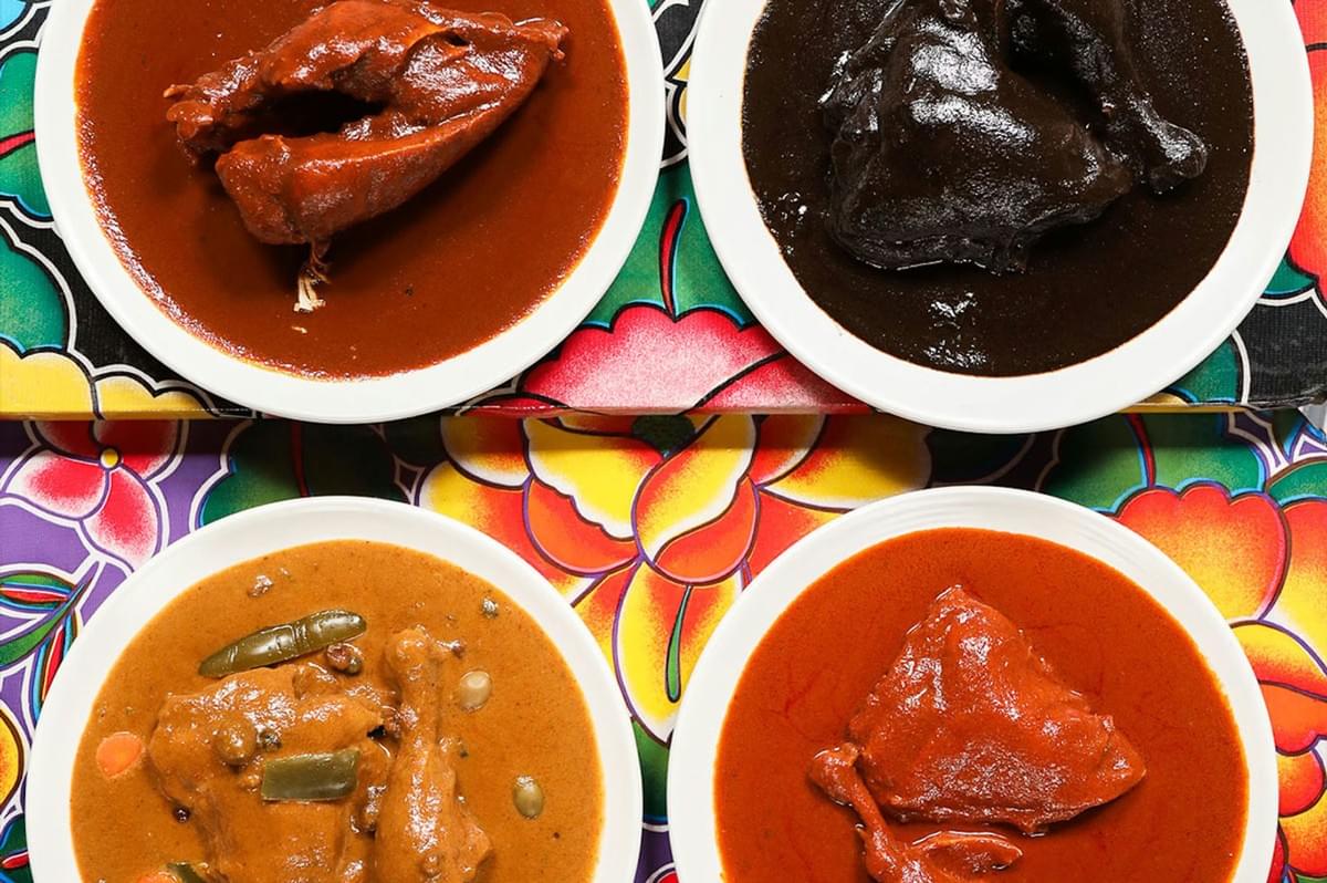 Overhead view of four plates, each containing a piece of chicken or meat covered in a different colored Mexican mole sauce: Mole Rojo, Mole Negro, Mole Verde, and Mole Poblano.