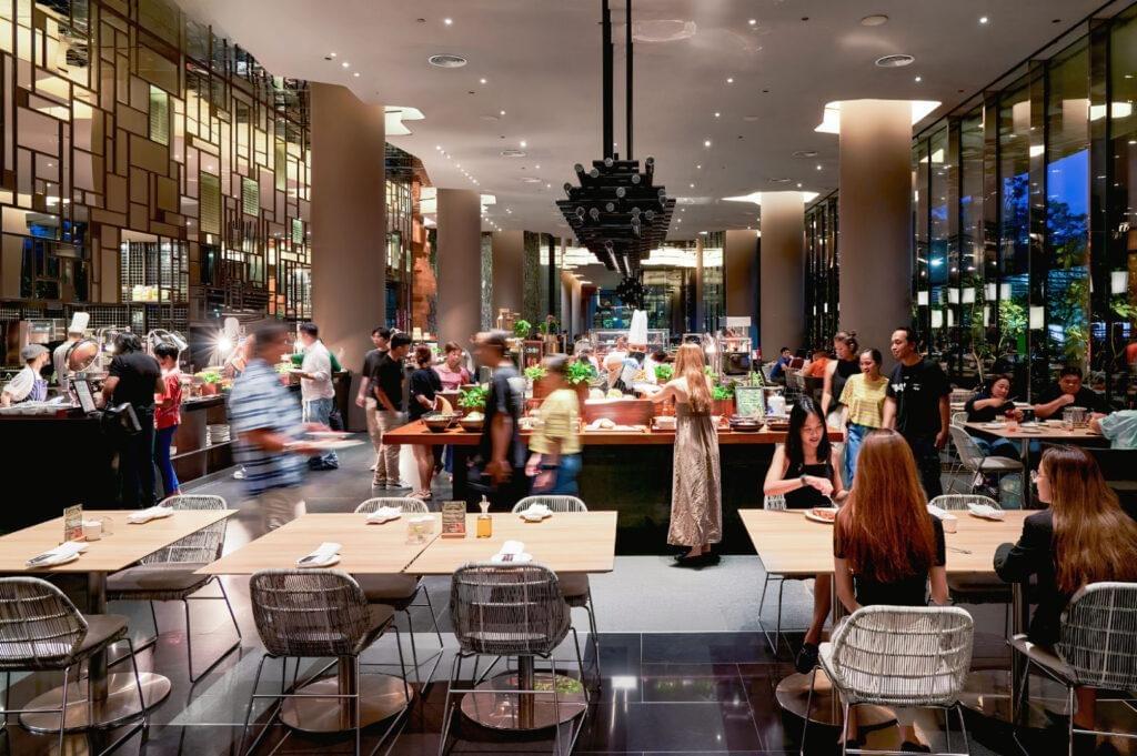 A spacious, modern restaurant interior features high ceilings, floor-to-ceiling windows, and elegant geometric wall paneling. Diners sit at wooden tables in the foreground while patrons browse food options at bustling buffet stations throughout the room.