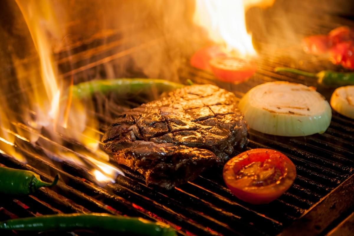 A thick, juicy steak sizzles over an open flame on a grill, surrounded by fresh vegetables like tomatoes, onions, and green peppers. The high-heat environment is captured through vibrant orange flares and rising smoke, highlighting the intense searing process of a barbecue.