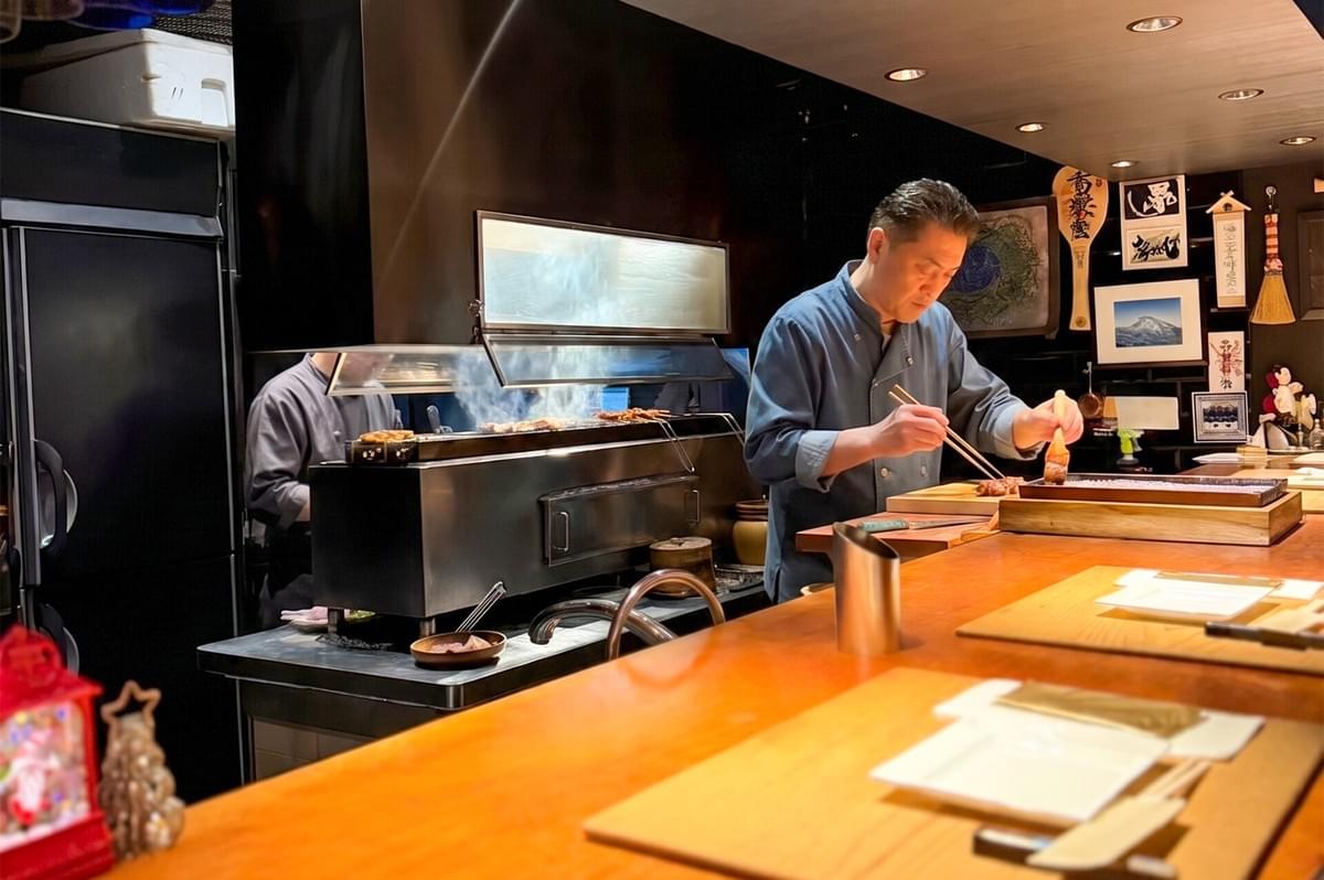 Professional chef preparing grilled skewers behind a wooden counter at Kazu Sumiyaki Restaurant.