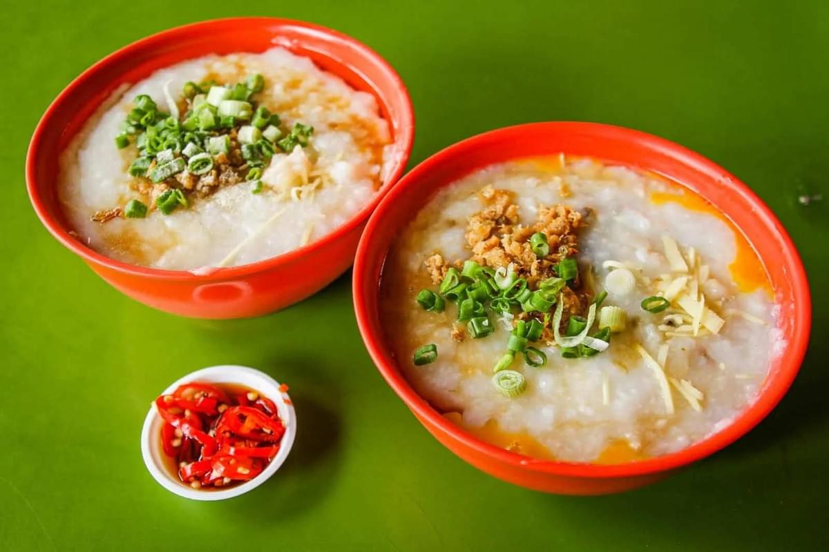 Two red bowls of thick, silky congee are topped with chopped spring onions, fried shallots, and ginger strips on a vibrant green table. A small side dish of sliced red chilies in soy sauce sits nearby, adding a sharp pop of color to the meal.