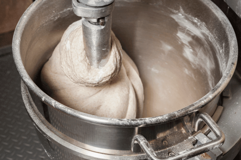 A mechanical spiral mixer kneading a large batch of pale bread dough in a stainless steel bowl, showing the initial stage of gluten bonding.