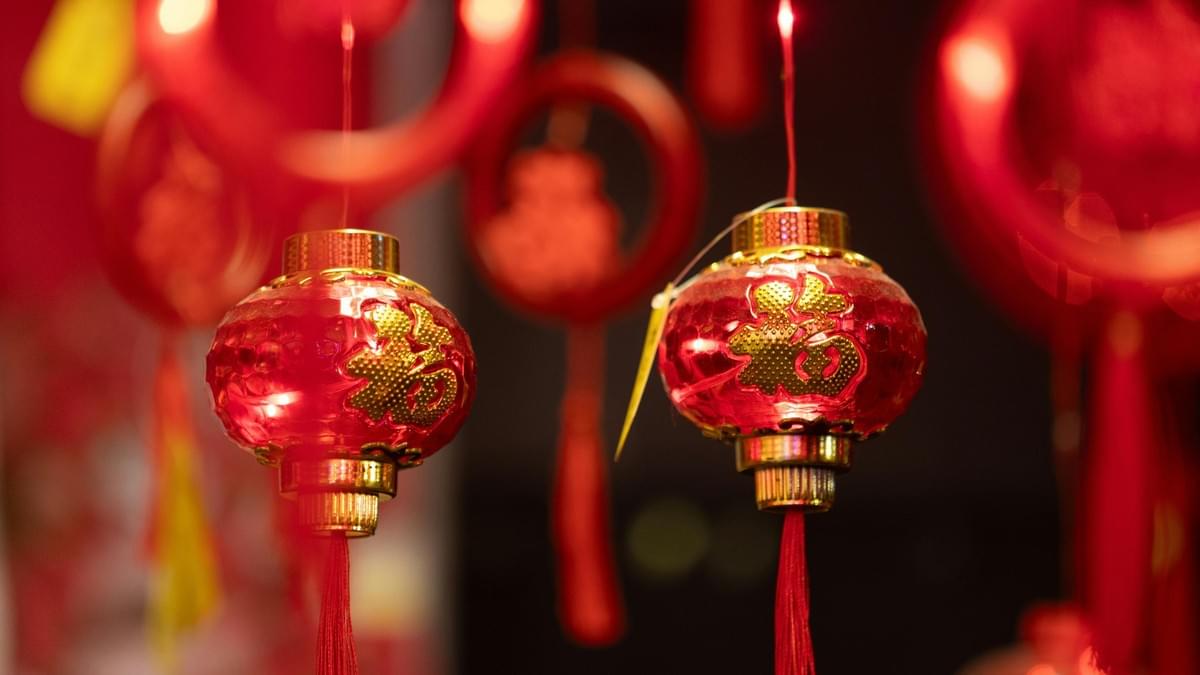 This image features a close-up of small, glossy red lanterns adorned with gold Chinese characters and long, flowing tassels. The blurred background of warm, glowing red tones creates a festive atmosphere, evoking the spirit of traditional Lunar New Year celebrations.