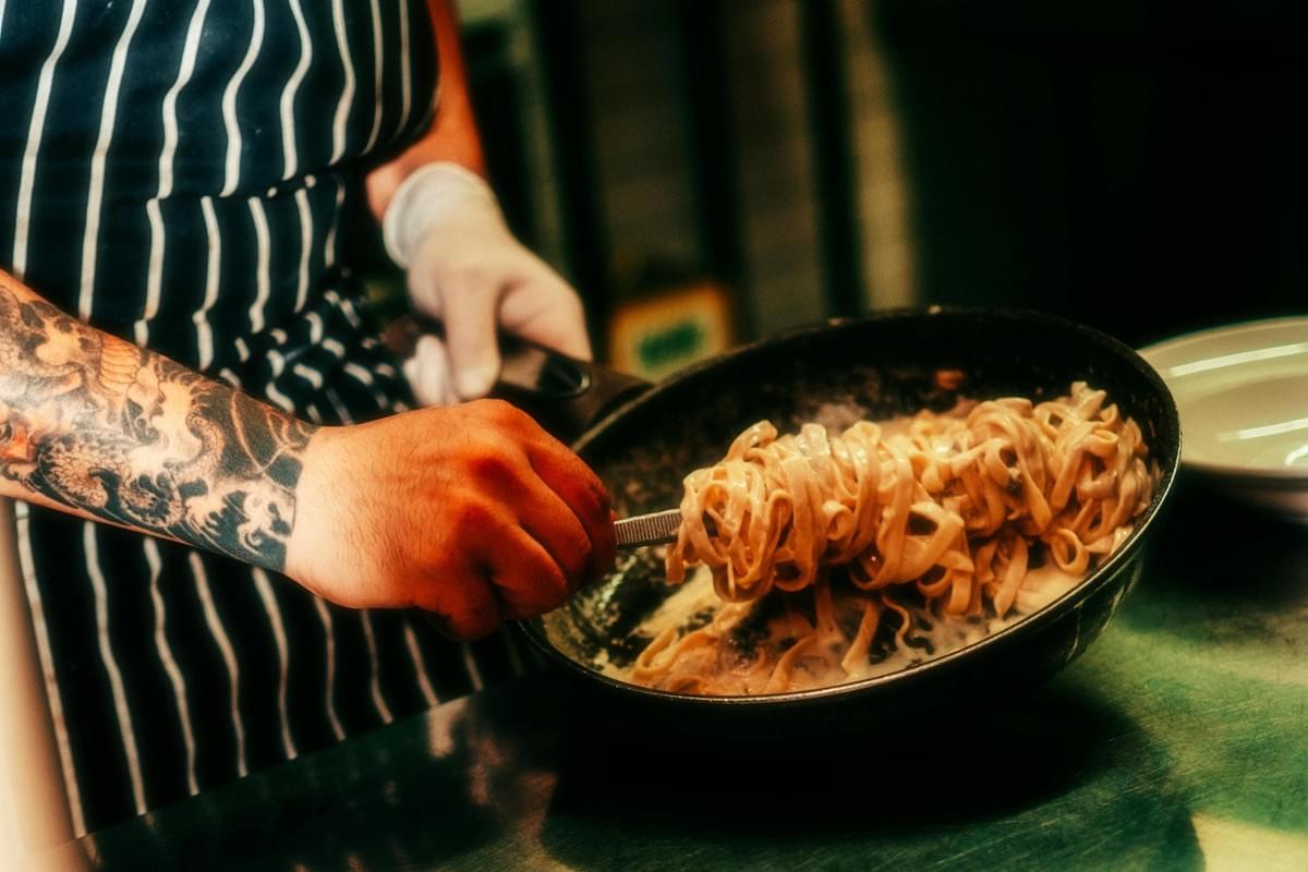 This close-up action shot features a chef with a heavily tattooed arm lifting a large serving of creamy pasta from a skillet using tongs. The warm lighting emphasizes the rich texture of the sauce-coated noodles and the chef's striped apron, capturing the dynamic energy of professional food preparation.