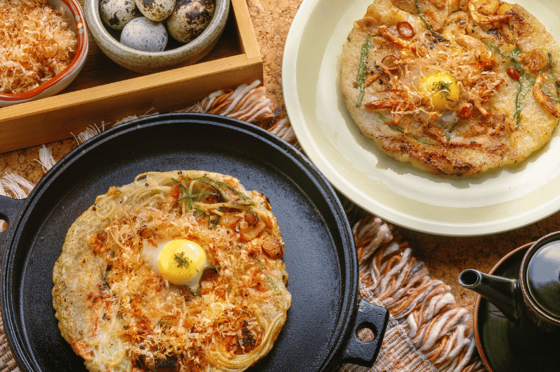 Two savory pancakes—one on a skillet, one on a plate—each crowned with a raw quail egg, vibrant vegetables, and fluttering bonito flakes, offering a bold, artful take on East Asian comfort food.
