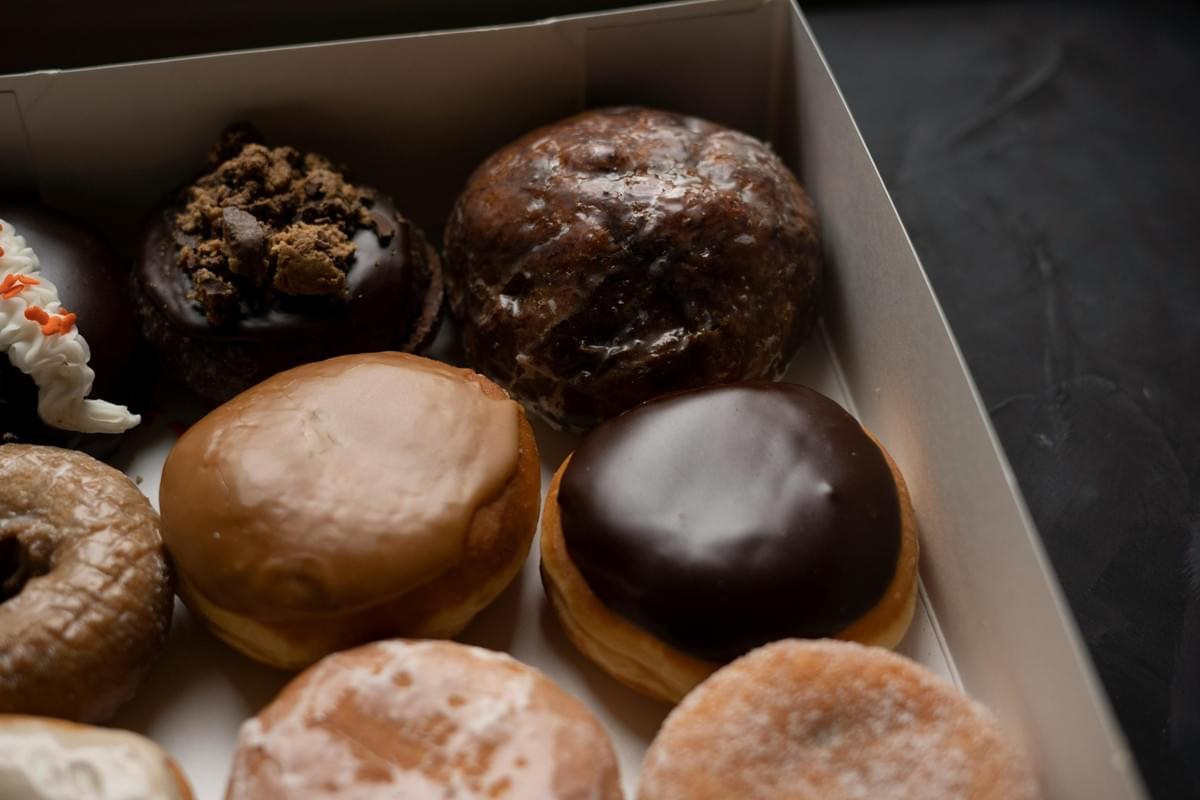 An open white bakery box is filled with an assortment of gourmet donuts featuring various toppings and glazes. The selection includes smooth chocolate-covered, maple-glazed, sugar-dusted, and chocolate-crumb-topped options set against a dark background.