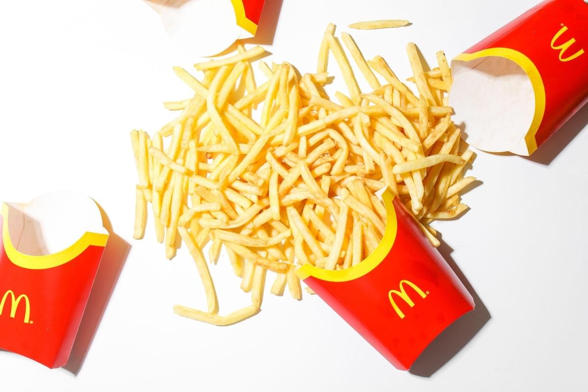 A top-down view shows a large pile of golden McDonald's french fries spilled across a bright white surface. Iconic red and yellow fry cartons surround the pile, with one box resting filled amidst the scattered fries.