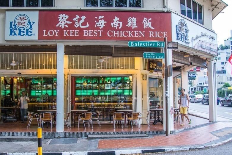  The image shows the exterior of Loy Kee Best Chicken Rice, a well-known eatery located on a corner along Balestier Road in Singapore. The storefront features a prominent red sign with white lettering, outdoor café-style seating under a covered walkway, and a green-backlit interior display.