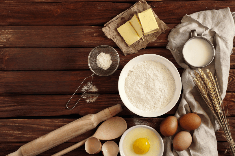 A rustic baking scene featuring flour, eggs, butter, milk, and utensils arranged on a wooden surface—evoking the warmth and anticipation of homemade cake preparation.