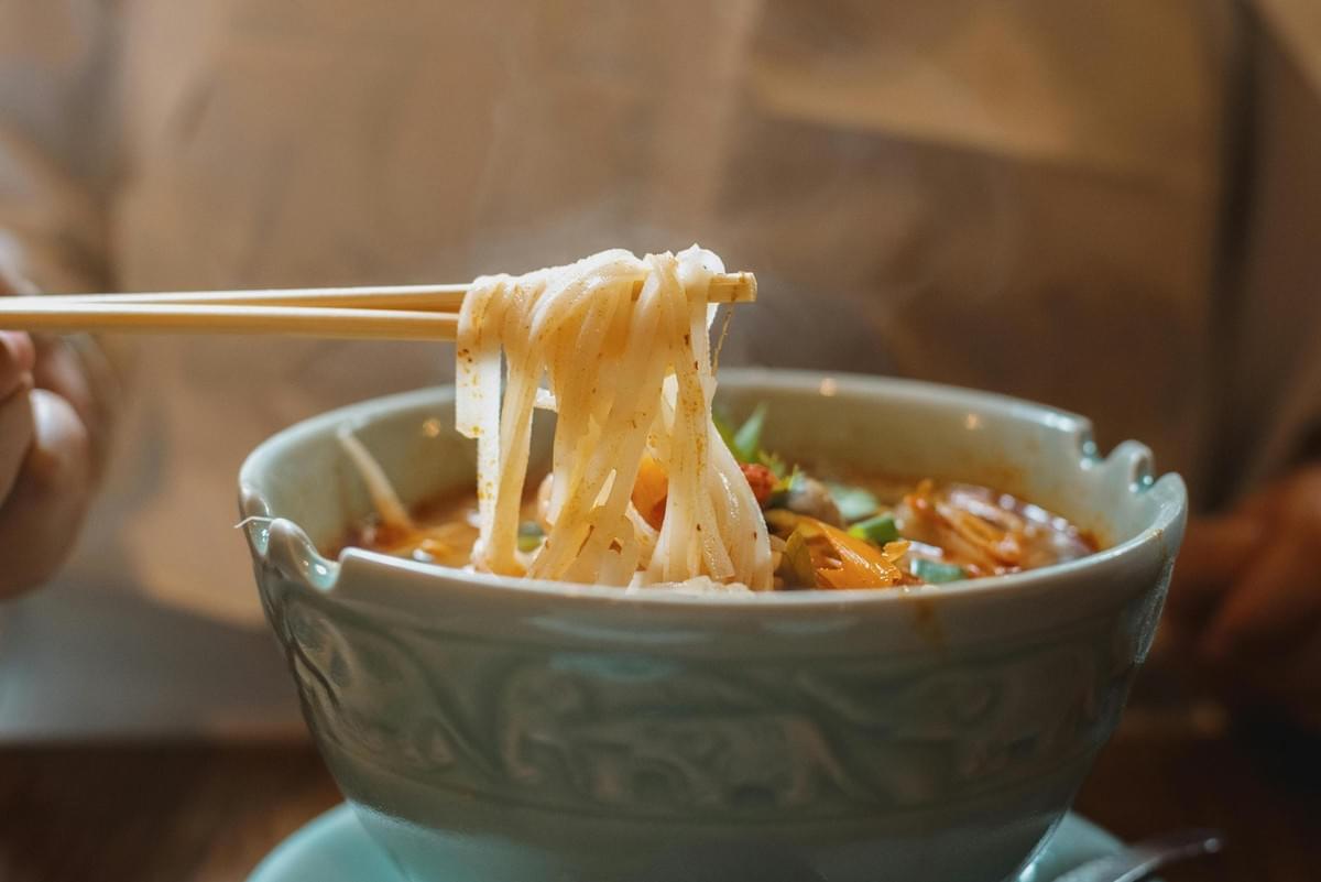 A female vendor stands amidst rising smoke at a street food stall packed with grilled delicacies, ranging from whole fish to various cuts of meat. Golden-brown roasted poultry hangs from hooks on the right, illuminated by a bright overhead bulb that casts a glow over the savory display.