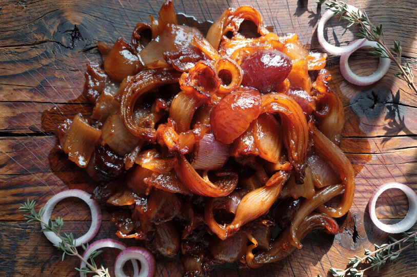 A top-down view of deeply caramelized red and yellow onions piled on a rustic wooden board, garnished with fresh thyme sprigs and raw red onion rings for contrast.
