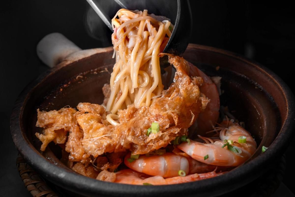 A steamy portion of Hokkien mee noodles is lifted with a spoon out of a rustic clay pot. The dish is loaded with fresh cooked prawns, crispy fried egg, and chopped green onions set against a dark background.