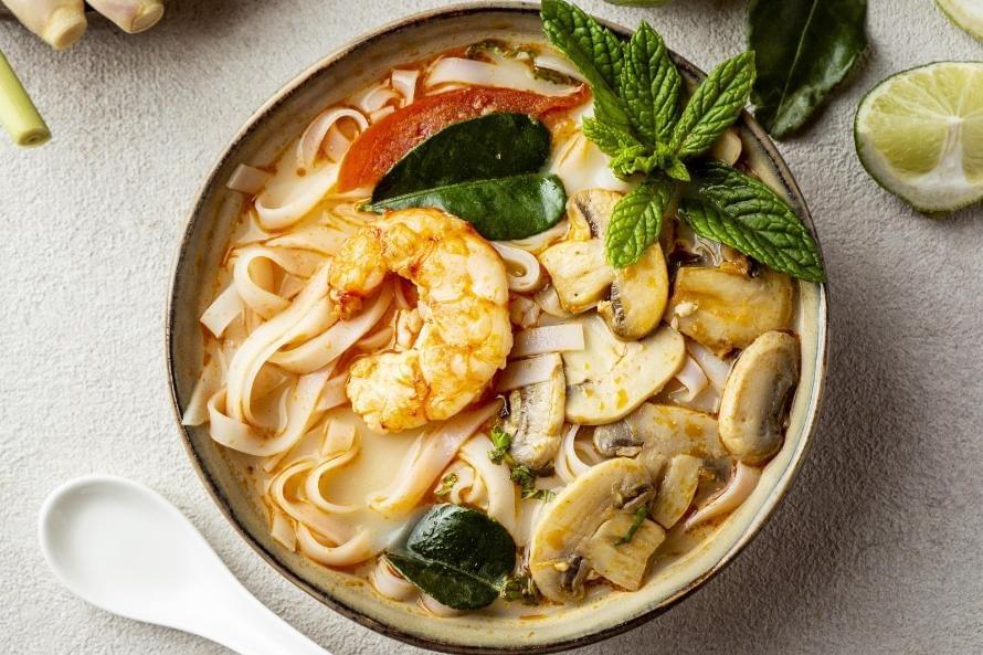 A close-up overhead shot a laksa bowl perfectly presented with all its ingredients with utensils and spices on the side.