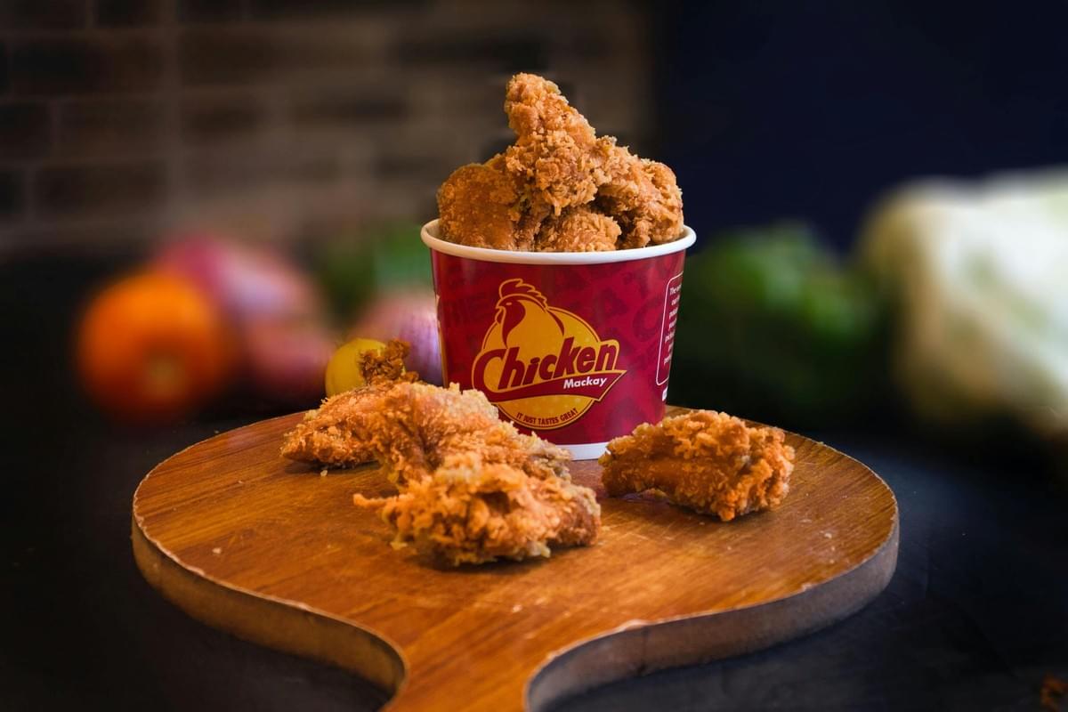 A red bucket filled with crispy, golden-fried chicken sits on a wooden cutting board with a handle. Several more pieces of fried chicken are scattered on the board in front of the bucket against a darkly lit, blurred background.