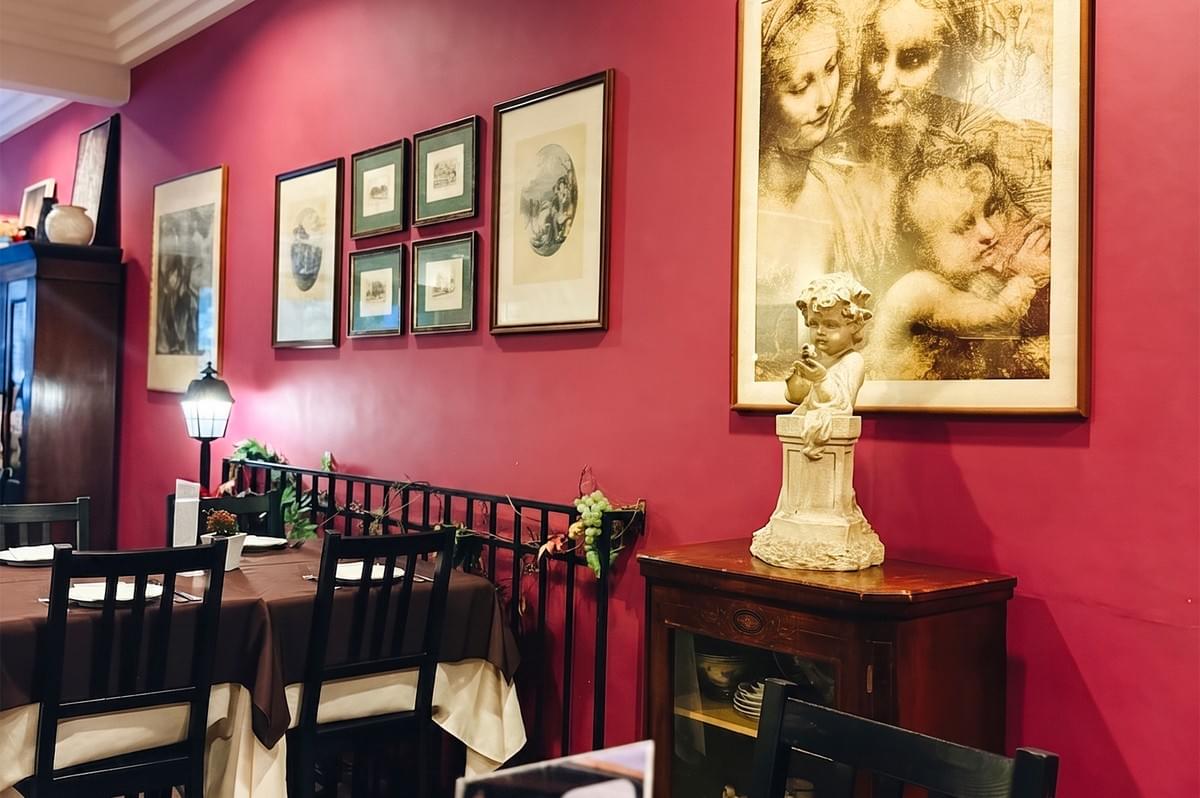 Cozy restaurant interior with deep red walls, framed artwork, and a cherub statue on a wooden cabinet near a dining table.