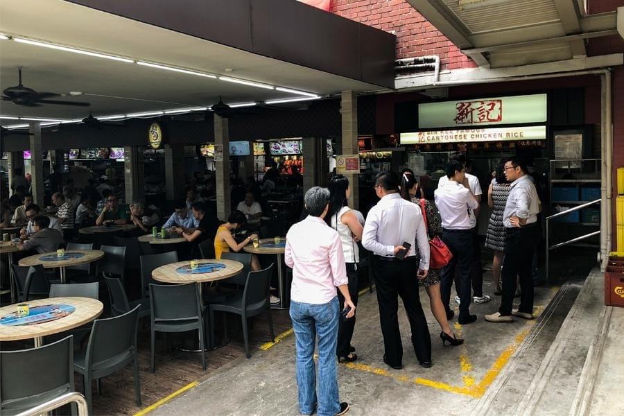  This image shows the Sin Kee Famous Cantonese Chicken Rice stall, featuring a glowing white signboard and a long queue of customers waiting for their food. The stall is located within a busy outdoor eating area filled with several round wooden tables and grey chairs, where many diners are already seated.