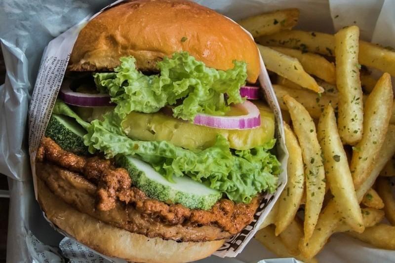 A large burger is stacked with a textured patty, fresh lettuce, red onion, and a slice of pineapple on a toasted bun. It is served in a paper-lined basket alongside a generous portion of seasoned, golden french fries.