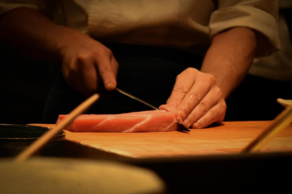 A sushi chef uses a sharp knife to slice a piece of fresh raw tuna on a wooden cutting board. The warm, focused lighting highlights the precision of the chef's hands during food preparation.
