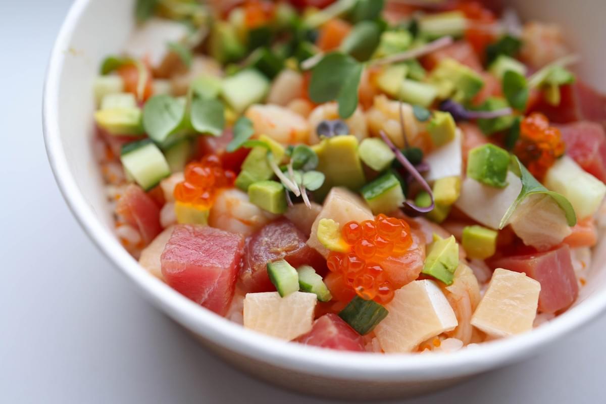 A white bowl filled with a colorful poke dish featuring diced raw tuna, salmon, white fish, and cucumber. Bright orange salmon roe, cubed avocado, and fresh microgreens garnish the top of the bowl.