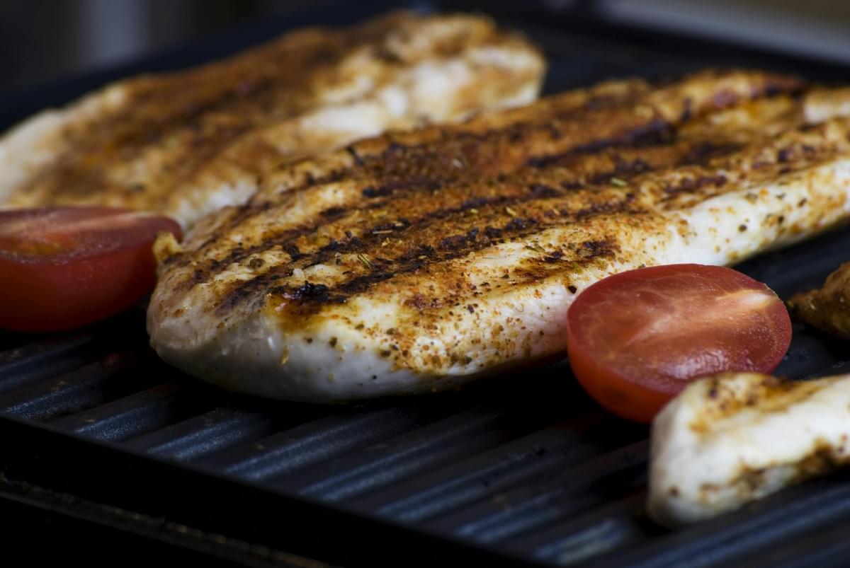 This close-up photograph focuses on a seasoned fillet of white meat, likely chicken or fish, being grilled on a black, ridged griddle, marked with distinct char lines.