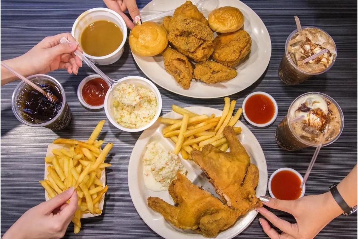 A top-down, flat-lay view captures a table spread with golden-brown fried chicken pieces, french fries, coleslaw, buns, gravy, and dipping sauces. Several hands reach into the frame to enjoy the meal alongside cups of iced cola and ice cream floats.
