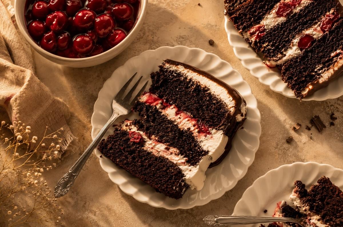 An overhead shot of a slice of layered chocolate cherry cake on a white ruffled plate with a fork, next to a bowl of glazed cherries.