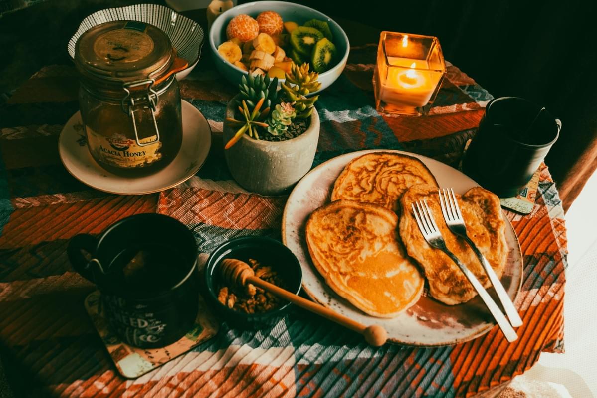 A cozy breakfast spread features a plate of golden pancakes with forks, accompanied by a jar of acacia honey, walnuts, and a bowl of mixed fruits. Two black mugs, a small potted succulent, and a lit candle sit on the patterned tablecloth, adding warmth to the inviting morning scene.