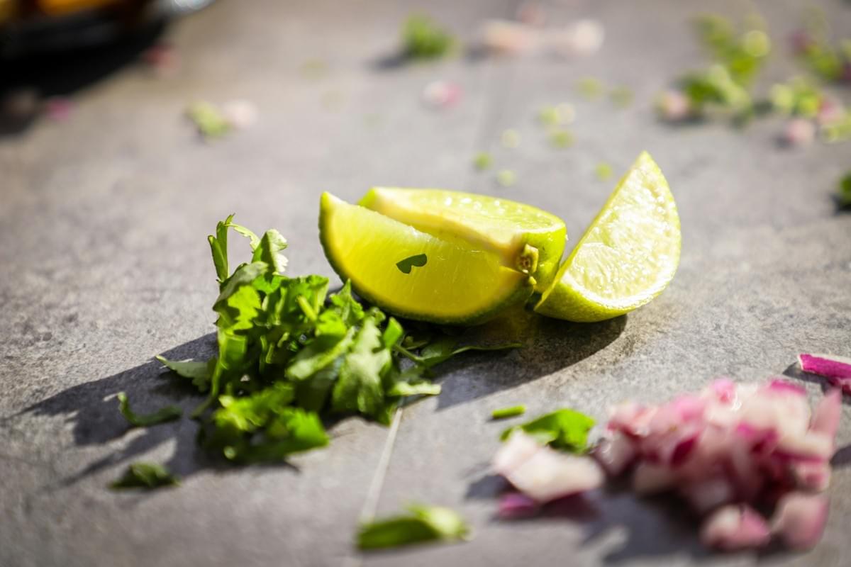 A close-up shot shows freshly chopped cilantro, lime wedges, and diced red onions scattered across a gray stone countertop. Bright sunlight casts distinct shadows, highlighting the vibrant green and purple colors of the taco ingredients.