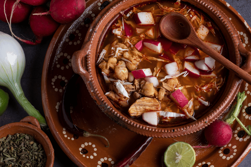 A top-down view of Pozole Rojo served in a traditional patterned clay pot, filled with tender pork, hominy, and chopped radishes, surrounded by whole onions and dried chilies.