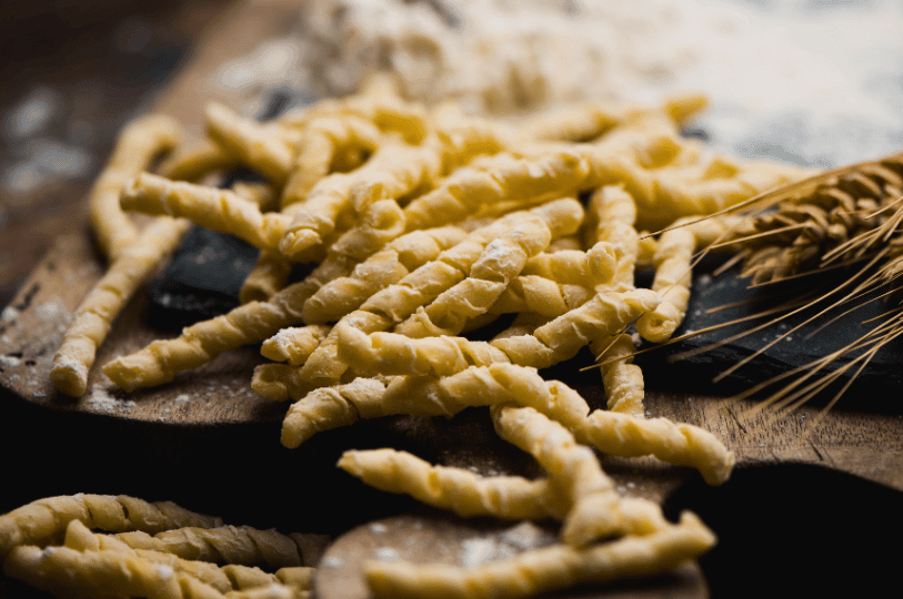 Flour-dusted, handmade twisted pasta, traditional Sicilian busiate, resting on a wooden board next to stalks of wheat.