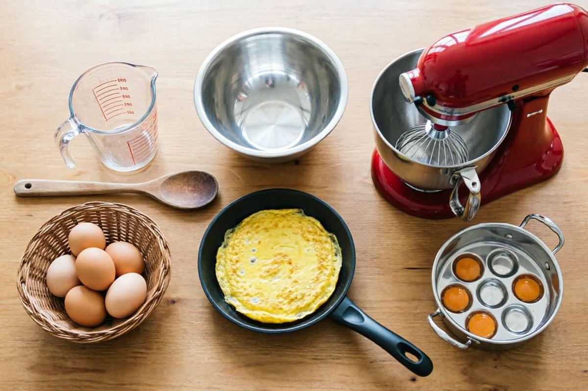 Egg cooking tools arranged on wooden table including mixer, pans, and ingredients