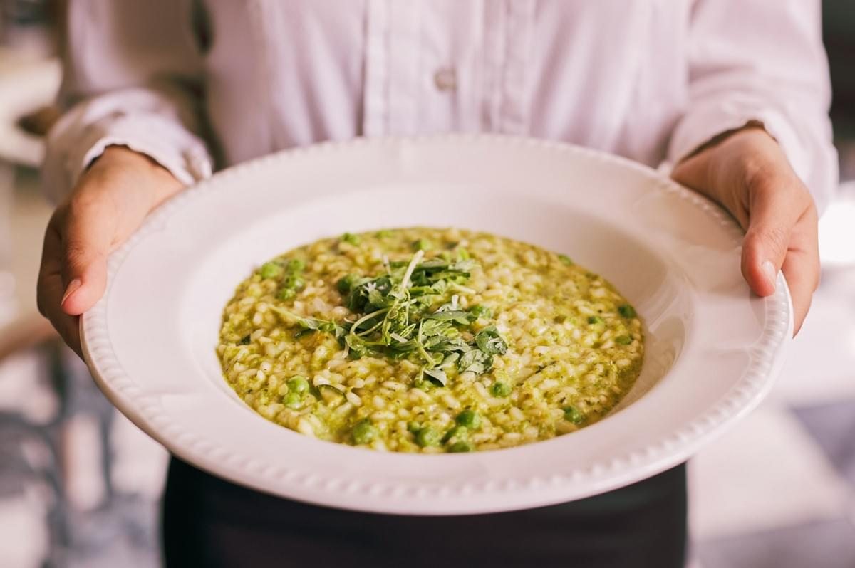 White plate filled with green herb risotto topped with fresh microgreens held over a restaurant table.