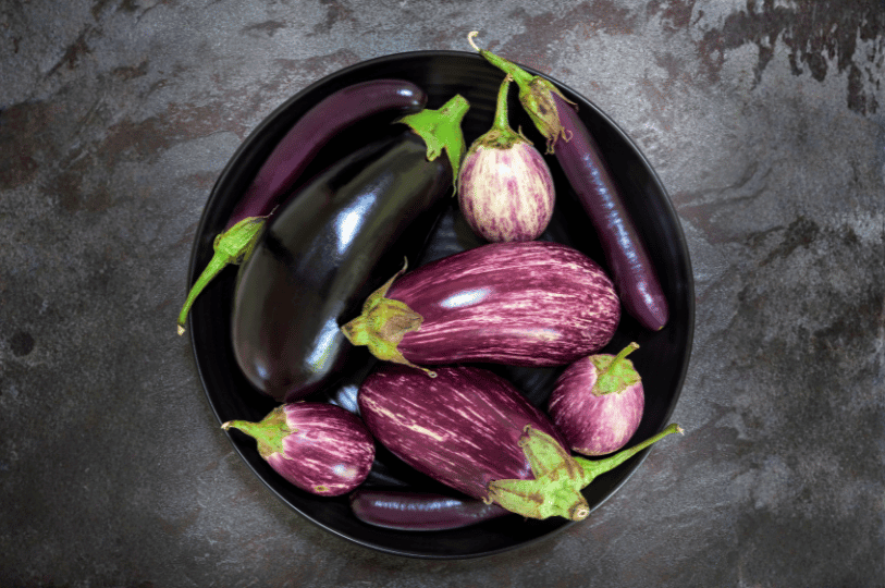 A black ceramic bowl containing a variety of eggplants, including a large Globe eggplant, slender Fairytale eggplants with white stripes, and small round Thai eggplants.