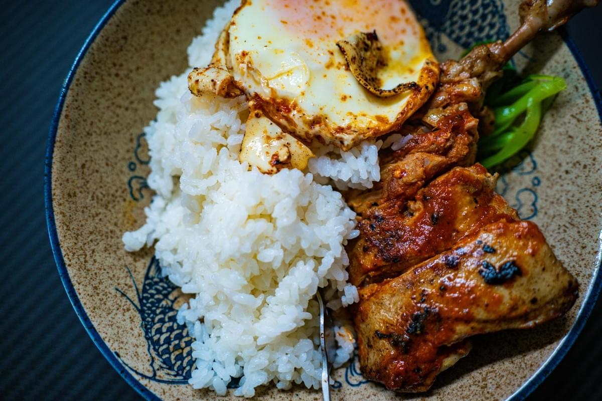 A bowl of white rice is topped with a fried egg and served alongside pieces of roasted chicken coated in a reddish sauce. The meal is presented in a ceramic bowl decorated with blue fish patterns and includes a side of bright green vegetables.