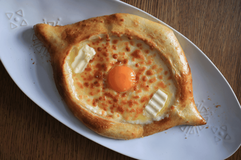 A top-down view of a golden-brown Khachapuri on a white oval plate. The boat-shaped bread is filled with melted, bubbling cheese, two thick pats of butter, and a bright orange raw egg yolk in the center.